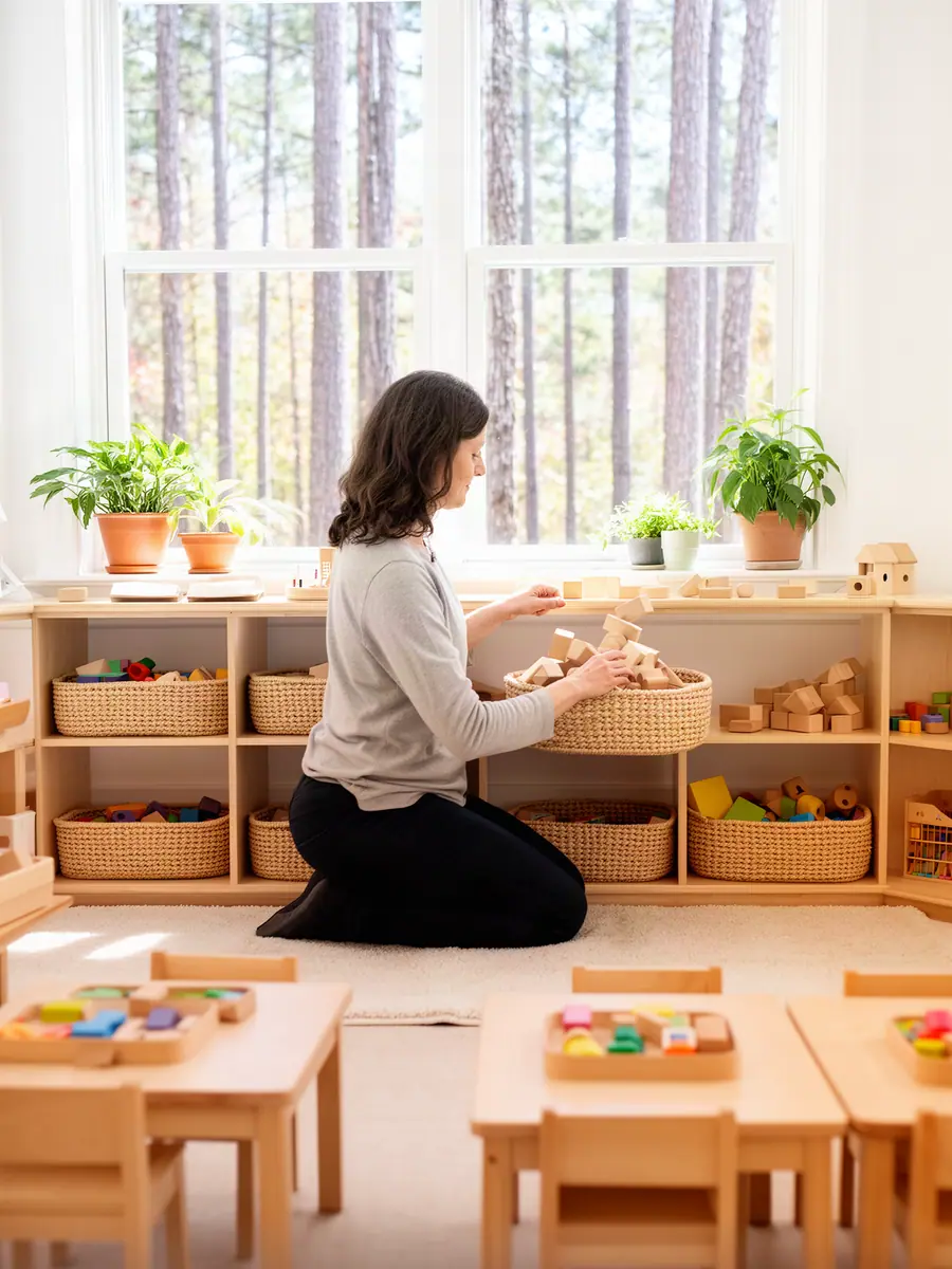 A child-care owner kneeling to set wooden toys and baskets on low shelves in a bright classroom before the children arrive.
